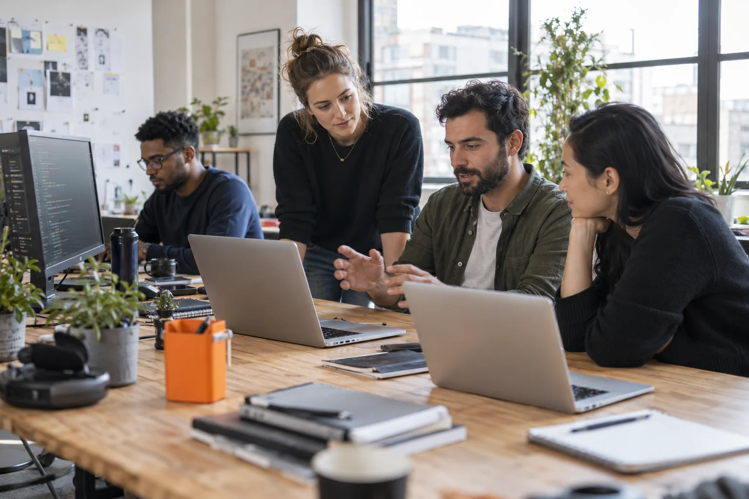 Software developers collaborating around laptops at a shared desk in a modern light-filled studio.